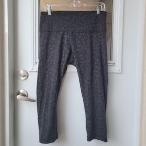 May include: A pair of dark charcoal gray leggings featuring a tonal, subtle leopard print pattern throughout. The leggings have a high-rise waistband and a cropped length that hits mid-calf, constructed with visible seams along the inner legs.