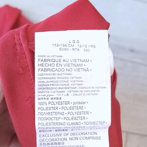 以下が含まれることがあります： 複数の言語でテキストが書かれた赤い衣料品タグのクローズアップ。タグには洗濯表示と素材構成が含まれており、ポリエステル100％と記載されています。タグには、衣料品がベトナム製で、12〜13歳向け、身長152〜158cmであることも記載されています。