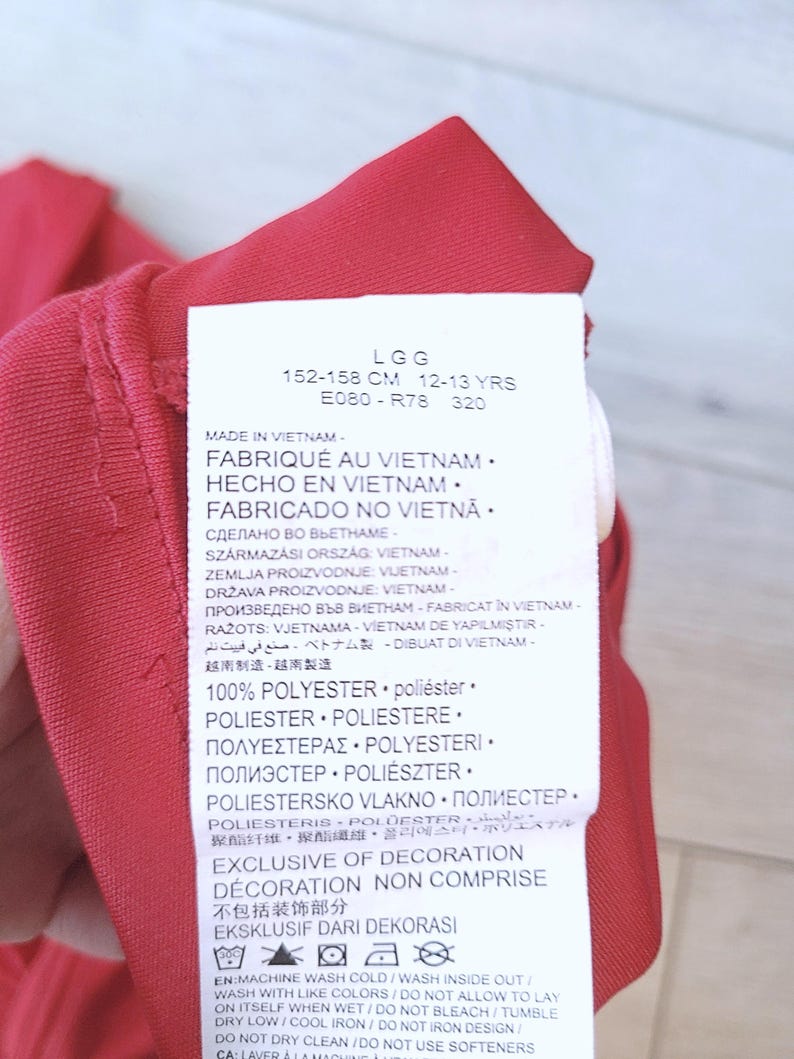以下が含まれることがあります： 複数の言語でテキストが書かれた赤い衣料品タグのクローズアップ。タグには洗濯表示と素材構成が含まれており、ポリエステル100％と記載されています。タグには、衣料品がベトナム製で、12〜13歳向け、身長152〜158cmであることも記載されています。