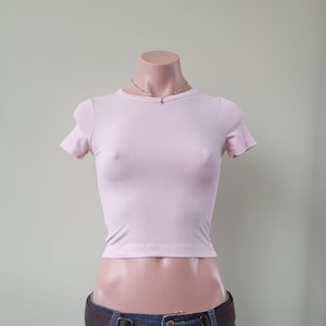 Könnte beinhalten: Ein hellrosa, kurzärmeliges, geripptes T-Shirt mit Rundhalsausschnitt. Das Kleidungsstück hat eine körpernahe Passform und ein strukturiertes, vertikales Rippenmuster im gesamten Stoff. Eine dünne silberfarbene Kette mit Anhänger wird getragen.