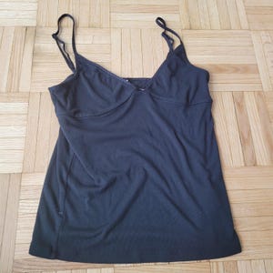 Peut inclure: Un caraco noir avec de fines bretelles spaghetti et un d&eacute;collet&eacute; en V. Le haut a un corsage ajust&eacute; et un bas l&eacute;g&egrave;rement &eacute;vas&eacute;. Le tissu semble &ecirc;tre un mat&eacute;riau tricot&eacute; doux. Le caraco est pos&eacute; sur une surface en bois.