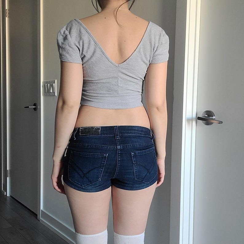 May include: A gray, short-sleeved crop top with a V-neck is paired with dark blue denim shorts. The shorts have a classic five-pocket design. White knee-high socks complete the outfit. The person is standing in front of a white door.