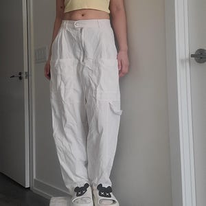 May include: Off-white cargo pants with multiple pockets and a zipper closure. The pants are paired with white slides featuring black bear face designs. The person is also wearing a yellow crop top.