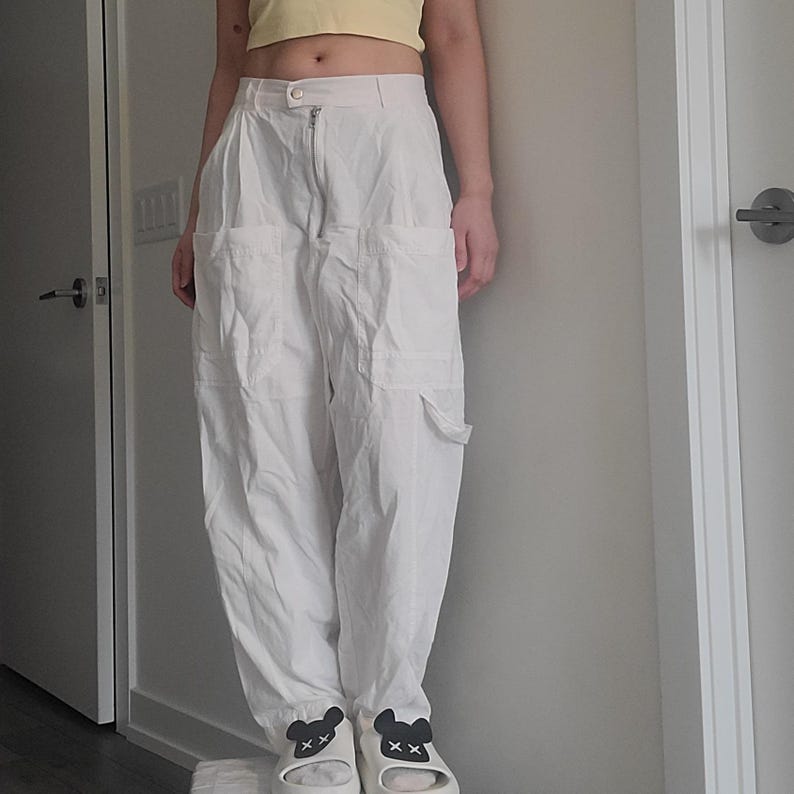 May include: Off-white cargo pants with multiple pockets and a zipper closure. The pants are paired with white slides featuring black bear face designs. The person is also wearing a yellow crop top.
