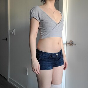 May include: A gray wrap-style crop top with short sleeves and a deep V-neck. Paired with dark blue denim shorts. The outfit is completed with white knee-high socks.