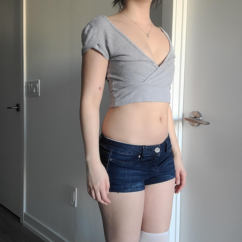 May include: A gray wrap-style crop top with short sleeves and a deep V-neck. Paired with dark blue denim shorts. The outfit is completed with white knee-high socks.
