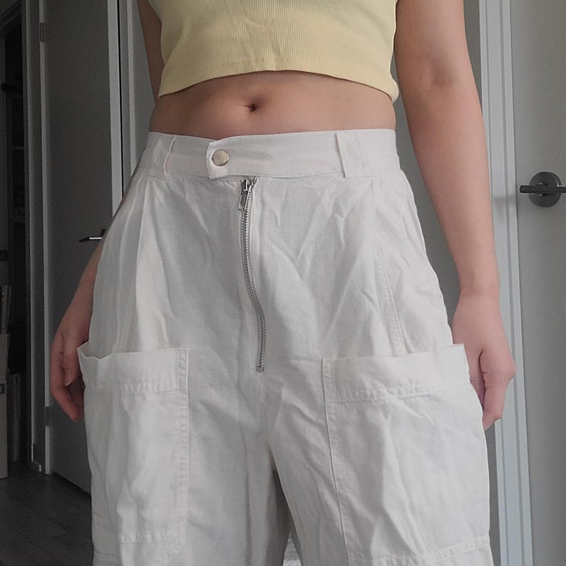 May include: Off-white, high-waisted pants with large front pockets and a silver zipper. The pants have a button closure at the waist and are made of a lightweight fabric. The pants are part of a casual outfit.