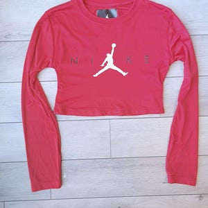 以下が含まれることがあります： 黒い文字で「NIKE」の文字とバスケットボール選手の白いシルエットが入った、赤い長袖クロップドトップ。クルーネックで、柔らかい素材で作られています。