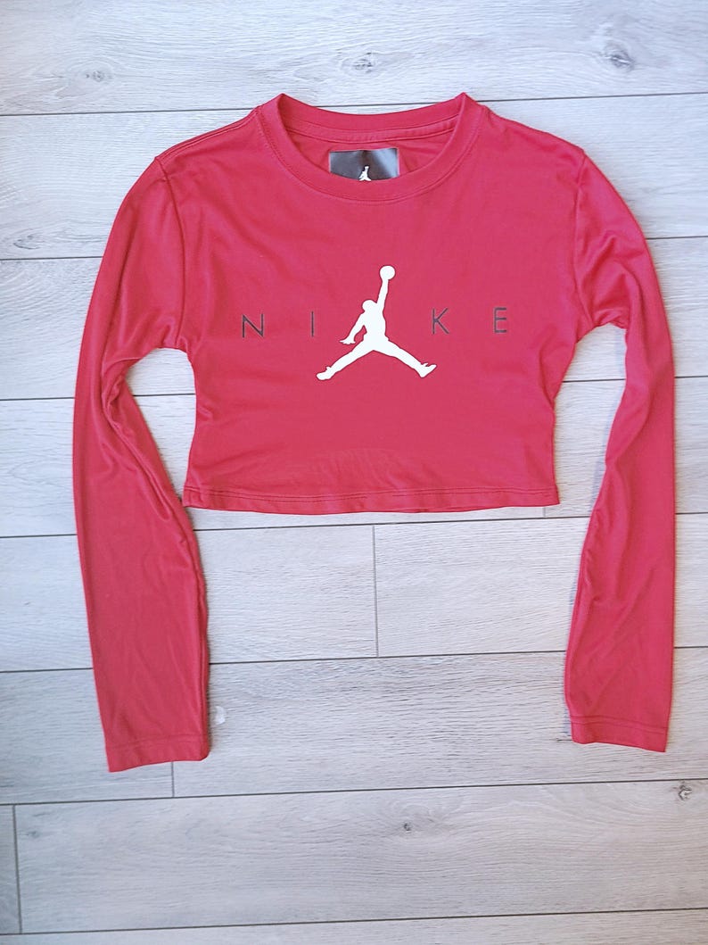 以下が含まれることがあります： 黒い文字で「NIKE」の文字とバスケットボール選手の白いシルエットが入った、赤い長袖クロップドトップ。クルーネックで、柔らかい素材で作られています。