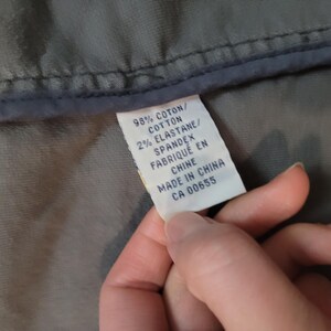Op de afbeelding: Close-up van een wit kledinglabel met zwarte tekst. Het label luidt "98% COTTON, 2% ELASTANE, SPANDEX, FABRIQU&Eacute; EN CHINE, MADE IN CHINA, CA 00655." Het label wordt tegen grijze stof gehouden.