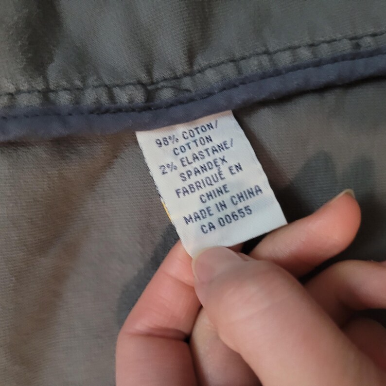 Op de afbeelding: Close-up van een wit kledinglabel met zwarte tekst. Het label luidt "98% COTTON, 2% ELASTANE, SPANDEX, FABRIQU&Eacute; EN CHINE, MADE IN CHINA, CA 00655." Het label wordt tegen grijze stof gehouden.