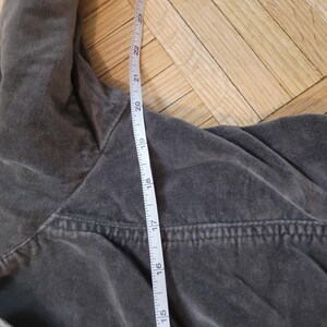 Op de afbeelding: Close-up van een grijs kledingstuk met een meetlint ernaast. De tape toont metingen in centimeters, waarbij de lengte van het kledingstuk wordt benadrukt. De stof lijkt een zacht, getextureerd materiaal te zijn.