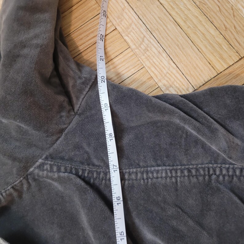 Op de afbeelding: Close-up van een grijs kledingstuk met een meetlint ernaast. De tape toont metingen in centimeters, waarbij de lengte van het kledingstuk wordt benadrukt. De stof lijkt een zacht, getextureerd materiaal te zijn.