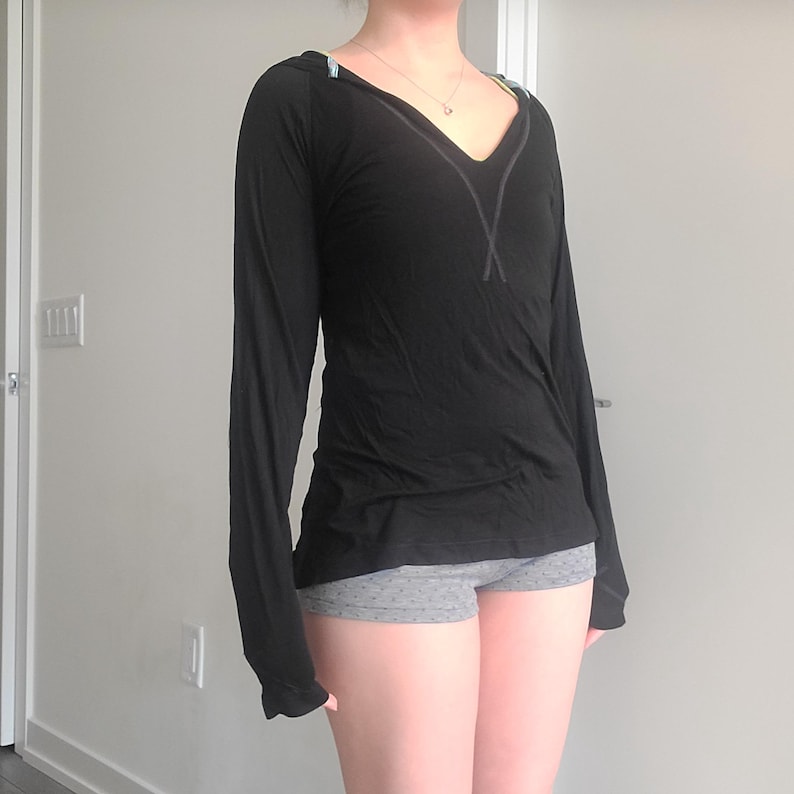 May include: Black long-sleeve shirt with a V-neck design and decorative stitching. The shirt has a relaxed fit and is paired with gray shorts. The shirt features a subtle design on the shoulders.