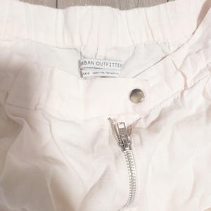 May include: Close-up of white shorts with a silver zipper and a snap closure. The shorts have an elastic waistband and a tag that reads "Urban Outfitters". The fabric appears to be lightweight.