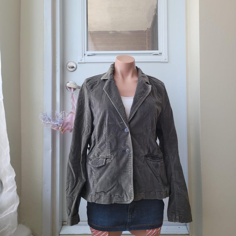 Op de afbeelding: Een grijze corduroy blazer met een reverskraag, twee voorzakken en een sluiting met &eacute;&eacute;n knoop. De blazer wordt getoond op een mannequin, gecombineerd met een spijkerbroekrok en een witte top.