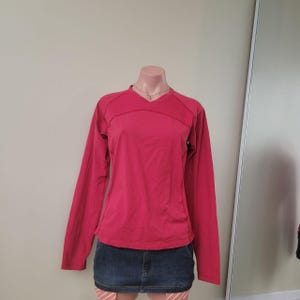 Peut inclure: Un haut à manches longues et col en V d'une couleur rose foncé unie. Le vêtement présente des coutures visibles sur la poitrine et le long des côtés, offrant une coupe structurée. Il est présenté sur un buste de mannequin avec une jupe en jean bleu.