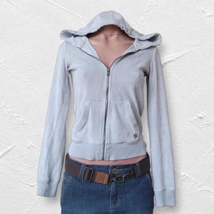 May include: A light blue zip-up hoodie featuring a hood, long sleeves with ribbed cuffs, and two front pockets. The garment is worn over a dark blue denim skirt secured with a brown belt that has a metal buckle and a brown leather accent piece.