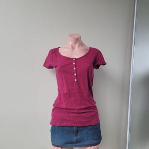 Puede incluir: Una camiseta henley de manga corta en color magenta oscuro con escote redondo y una tapeta vertical con cinco pequeños botones blancos en el centro. La prenda se combina con una minifalda de mezclilla azul con costuras visibles.