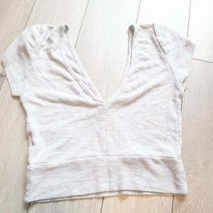 May include: A light beige crop top with a deep V-neckline and short sleeves. The top has a ribbed texture and a wide, banded hem. The fabric appears to be a soft, lightweight material, suitable for warm weather.