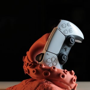 May include: A white and black video game controller held by an orange octopus-shaped stand. The controller has multiple buttons and a touchpad. The stand has a textured surface and suction cups.