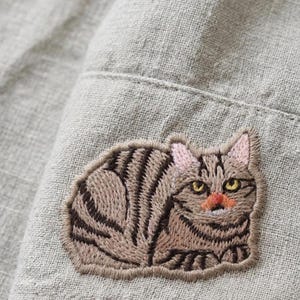May include: Close-up of a gray fabric with a detailed embroidered patch of a tabby cat. The cat has brown and black stripes, pink ears, and a red nose. The patch is sewn onto the fabric.