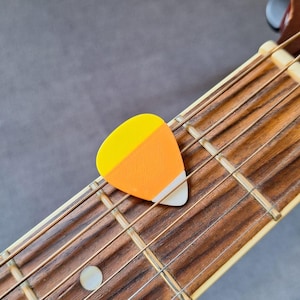 May include: A candy corn-shaped guitar plectrum sits on the fretboard of an acoustic guitar. The plectrum has yellow, orange, and white sections. The guitar's wooden fretboard and strings are visible, creating a musical and festive image.