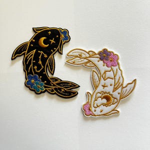 May include: Two embroidered patches shaped like koi fish. One patch is black with gold celestial patterns, a crescent moon, and blue flowers. The other patch is white with gold celestial patterns, a crescent moon, and pink and purple flowers.