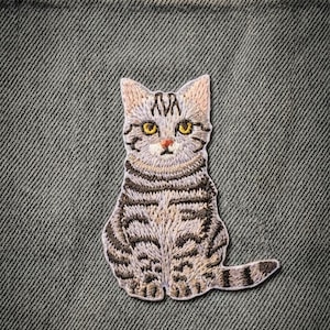 May include: Embroidered patch of a seated tabby cat with brown and grey stripes and yellow eyes. The patch is sewn onto a textured, grey fabric background. The cat's ears are a light tan colour.