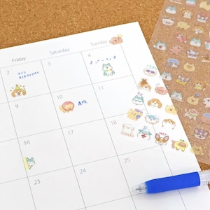 May include: A white calendar page with cartoon dog stickers and a blue pen. The calendar has dates and days of the week labeled. Some dates have Japanese text and stickers. A sheet of dog stickers is visible on the right.