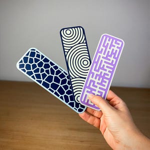 May include: Three decorative bookmarks with geometric patterns. One bookmark has a blue and white honeycomb design, another has concentric circles, and the third has a maze-like pattern. The bookmarks are held in a hand.