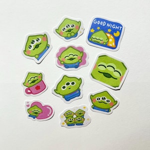 May include: A collection of ten die-cut stickers featuring a small green three-eyed alien character in various poses and settings, including one square sticker with the text GOOD NIGHT, a pink heart, a pink mug, and a group of four aliens.