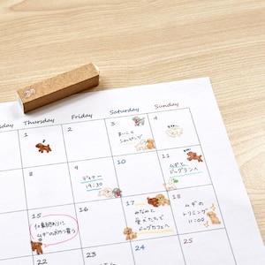 Pode incluir: Um calendário com ilustrações de cães e texto japonês, com um carimbo de madeira com uma imagem de cão. O calendário tem notas manuscritas e adesivos com tema de cães. O calendário está em uma superfície de madeira clara.