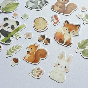 May include: A collection of woodland animal stickers, including a panda with bamboo, a fox, a squirrel, and a rabbit. Other stickers include acorns, flowers, leaves, and a mushroom. The stickers are in soft watercolor tones.