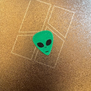 Könnte beinhalten: Ein grüner, außerirdisch geformter Gitarrenplektrum mit schwarzen Augen und einem kleinen Mund. Der Plektrum liegt auf einer strukturierten, goldfarbenen Oberfläche mit einem geometrischen Muster.