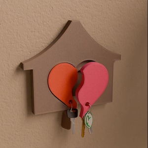 May include: A brown house-shaped key holder featuring a broken heart design. The heart is split into two halves, one orange and one pink. Keys hang from each heart half. The key holder is mounted on a beige wall.