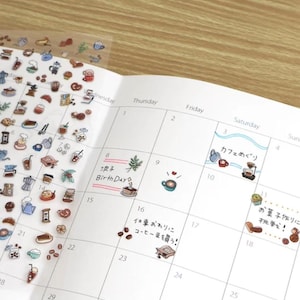 May include: A transparent sticker sheet featuring numerous small, colorful illustrations of coffee-related items, pastries, and drinks, partially overlapping a white paper calendar with handwritten Japanese text and small sticker accents.