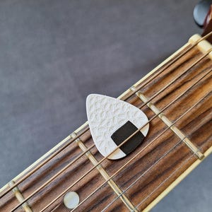 May include: A white guitar pick with a textured pattern and a black rectangular design, resting on the fretboard of a wooden acoustic guitar. The strings and frets are visible, with a gray background.