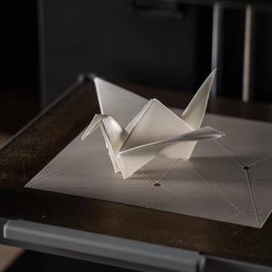 以下が含まれることがあります： 白い折り紙の鶴が3Dプリンターのベッドの上に置かれています。鶴は一枚の紙から折りたたまれ、幾何学模様の上に配置されています。プリンターは黒色で、金属的な外観をしています。