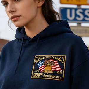 May include: A navy blue hooded sweatshirt featuring a rectangular embroidered patch on the chest. The patch displays the Liberty Bell, an American flag, and the text United States of America, 1776, 2026, and 250th Anniversary in gold.