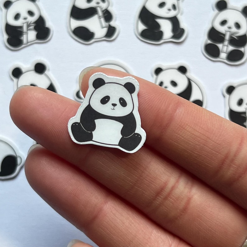 May include: A small die-cut sticker featuring a black and white illustration of a seated panda bear. The panda has a round face, black ears, black eye patches, and black limbs, with a white belly and face. The sticker has a thin white border.