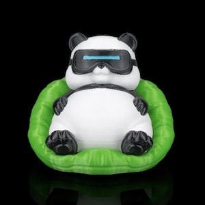 May include: A white panda figurine with black ears, eyes, and paws, wearing a black and blue VR headset. The panda is seated in a bright green, leaf-shaped cushion against a black background.