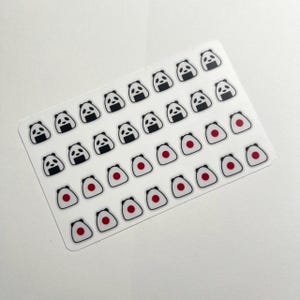 May include: A rectangular sticker sheet featuring multiple small, individual stickers arranged in rows. The designs include black and white panda faces and white rice balls with a central red circle, all outlined in a thin black border.