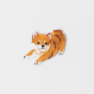 May include: Embroidered patch featuring a Shiba Inu dog in a playful pose. The patch showcases the dog's orange and white fur with detailed stitching. The dog is depicted with a black nose and eyes, and white paws.