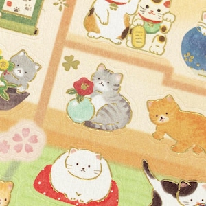 May include: A sheet of illustrated cat stickers in various poses and colours. The stickers feature cats with gold accents, some holding flowers or sitting on cushions. The background is a light beige with floral and geometric patterns.