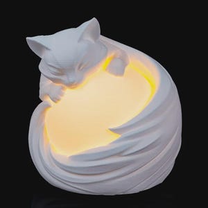 May include: A white cat-shaped lamp with a soft, glowing yellow light emanating from within. The cat is curled up, with its head resting on its paws. The lamp has a smooth, textured surface and is set against a dark background.