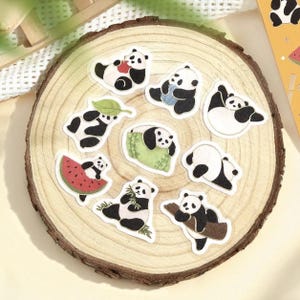 May include: A selection of panda bear stickers in different poses, such as eating bamboo, resting on a leaf, and lounging on a watermelon slice. The stickers are black and white with a white border, displayed on a wooden slice.