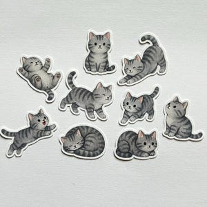 May include: A collection of nine gray tabby cat stickers in various poses. The stickers feature smiling cats in playful positions, including lying down, sitting, and stretching. The stickers are on a white background.