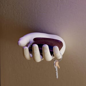 May include: A white hand-shaped wall hook holding a dark brown object and a set of keys. The hook is mounted on a neutral-toned wall. The keys are attached to a silver key ring.
