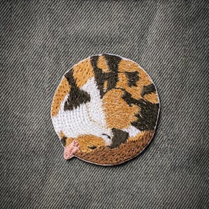 May include: Embroidered patch featuring a sleeping calico cat in a curled-up position. The patch is circular, with the cat's fur rendered in shades of brown, black, and white. The cat's nose is pink, and the patch is on a textured, gray background.
